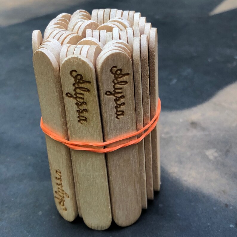 May include: A bundle of wooden craft sticks with the name "Alyssa" engraved on each stick. The sticks are tied together with a rubber band.