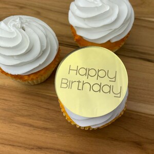 Może przedstawiać: Złoty topper do babeczek z napisem "Happy Birthday" na białej babeczce z białym lukrem.