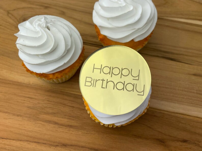 Może przedstawiać: Złoty topper do babeczek z napisem "Happy Birthday" na białej babeczce z białym lukrem.