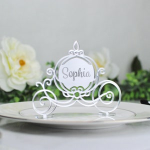 May include: White Cinderella carriage place card holder with the name "Sophia" written on it. The holder is sitting on a white plate.
