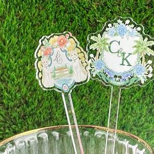 Puede incluir: Dos toppers de pastel de acr&iacute;lico transparente con dise&ntilde;os florales. El primer topper presenta una casa blanca con dos gatos y el texto "L&K". El segundo topper presenta un escudo con el texto "C&K" rodeado de palmeras y flores azules.
