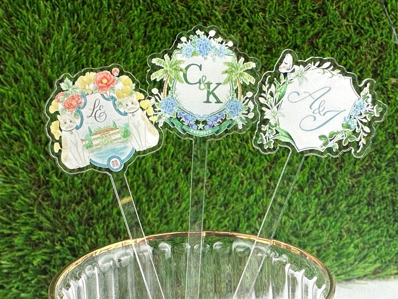 Puede incluir: Tres toppers de pastel de acr&iacute;lico transparente con dise&ntilde;os florales e iniciales personalizadas. Los toppers presentan un gato blanco, una palmera y una mariposa.