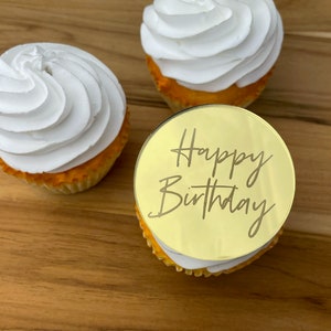 Może przedstawiać: Trzy białe, lukrowane babeczki z okrągłym, złotym topperem, na kt&oacute;rym napisano "Happy Birthday" ozdobnym pismem.