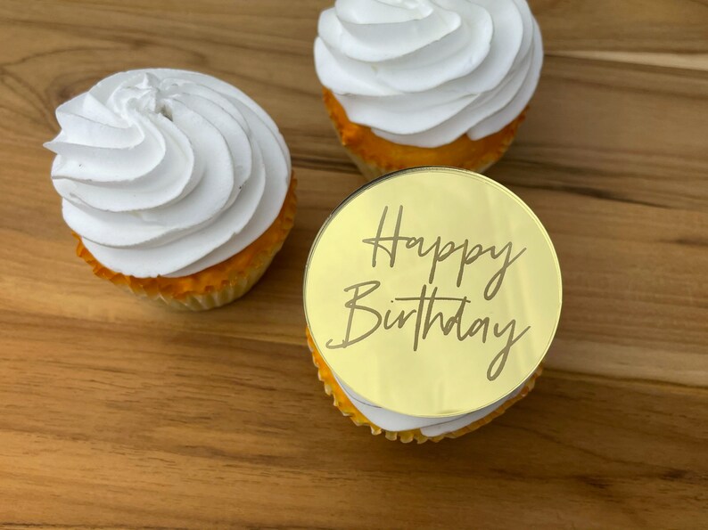 Może przedstawiać: Trzy białe, lukrowane babeczki z okrągłym, złotym topperem, na kt&oacute;rym napisano "Happy Birthday" ozdobnym pismem.