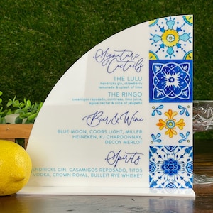 May include: White sign with blue and yellow tile pattern. The sign lists signature cocktails, beer and wine, and spirits. The cocktails listed are The Lulu and The Ringo. The beer and wine list includes Blue Moon, Coors Light, Miller, Heineken, KJ Chardonnay, and Decoy Merlot. The spirits list includes Hendricks Gin, Casamigos Reposado, Tito's Vodka, Crown Royal, and Bulleit Rye Whiskey.