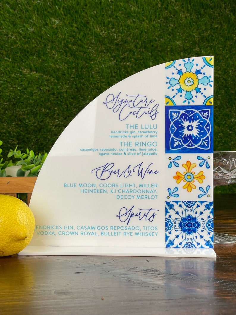 May include: White sign with blue and yellow tile pattern. The sign lists signature cocktails, beer and wine, and spirits. The cocktails listed are The Lulu and The Ringo. The beer and wine list includes Blue Moon, Coors Light, Miller, Heineken, KJ Chardonnay, and Decoy Merlot. The spirits list includes Hendricks Gin, Casamigos Reposado, Tito's Vodka, Crown Royal, and Bulleit Rye Whiskey.