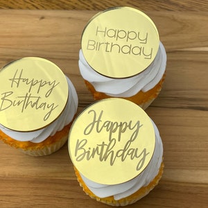 Może przedstawiać: Trzy złote babeczki z białym kremem i okrągłymi złotymi nakładkami lustrzanymi, na kt&oacute;rych napisano "Happy Birthday" r&oacute;żnymi czcionkami.