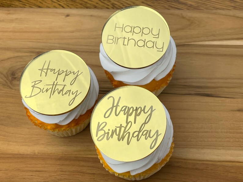 Może przedstawiać: Trzy złote babeczki z białym kremem i okrągłymi złotymi nakładkami lustrzanymi, na kt&oacute;rych napisano "Happy Birthday" r&oacute;żnymi czcionkami.