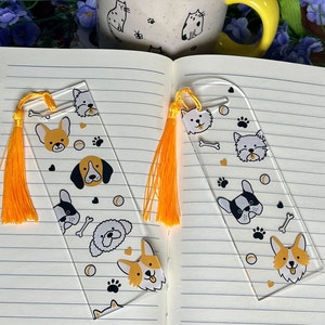 May include: Two clear acrylic bookmarks with yellow tassels. Each bookmark features a different design of various dog breeds, including a corgi, a bulldog, a poodle, and a beagle. The designs are in black and white with yellow accents.