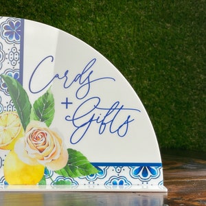 May include: A white ceramic sign with a blue and white patterned border. The sign is shaped like a half circle and has the text "Cards + Gifts" written in blue cursive script. The sign is decorated with a watercolor illustration of lemons, leaves, and a rose.