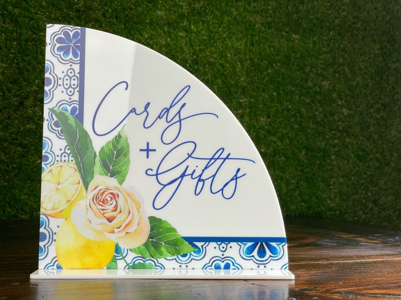 May include: A white ceramic sign with a blue and white patterned border. The sign is shaped like a half circle and has the text "Cards + Gifts" written in blue cursive script. The sign is decorated with a watercolor illustration of lemons, leaves, and a rose.