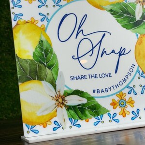 May include: A white sign with a blue circle and the text "Oh Snap" in blue cursive writing. Below the circle is the text "Share the Love" and the hashtag "#BABYTHOMPSON". The sign is decorated with a watercolor design of lemons, leaves, and flowers.