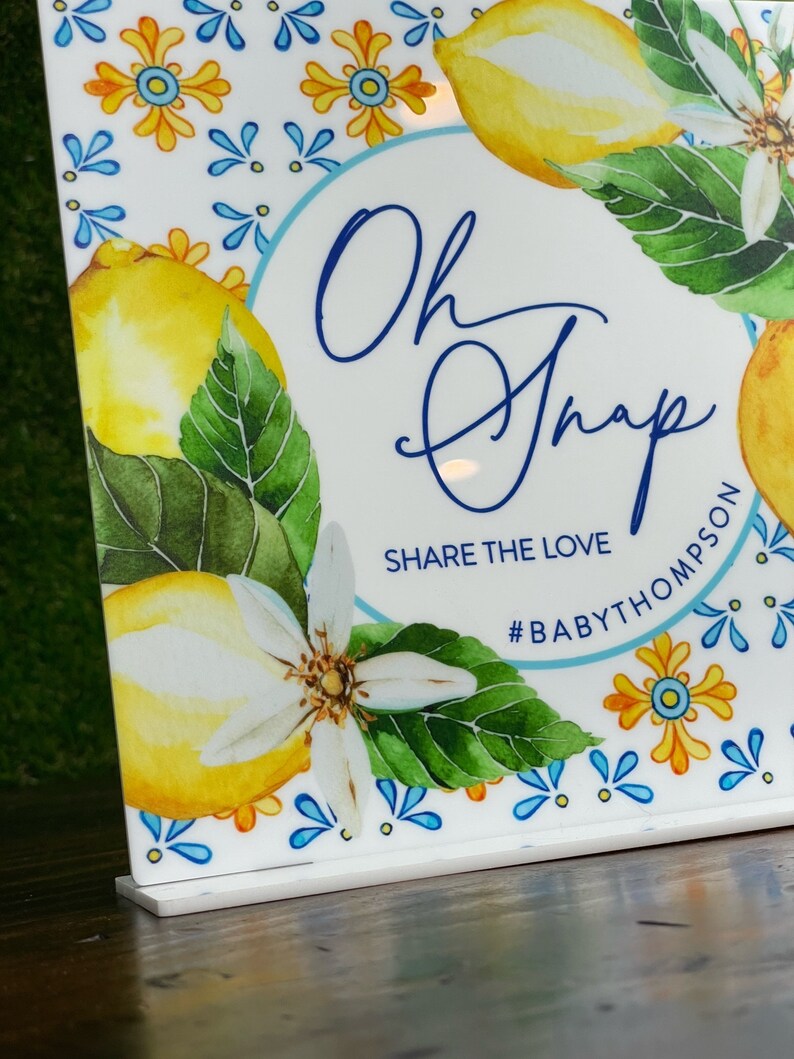 May include: A white sign with a blue circle and the text "Oh Snap" in blue cursive writing. Below the circle is the text "Share the Love" and the hashtag "#BABYTHOMPSON". The sign is decorated with a watercolor design of lemons, leaves, and flowers.