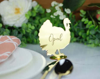 May include: Gold mirror acrylic turkey place card with the name "Opal" engraved on it. The place card is sitting on a white table with a plate and napkin.