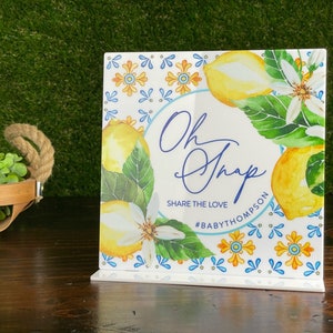 May include: A white square sign with a lemon and floral design. The sign says "Oh Snap" and "Share the Love #BabyThompson".