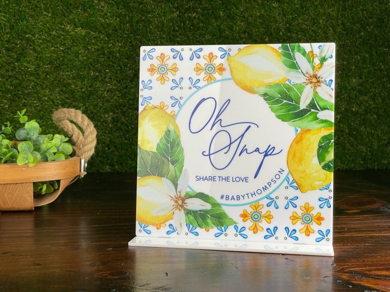 May include: A white square sign with a lemon and floral design. The sign says "Oh Snap" and "Share the Love #BabyThompson".