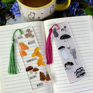 May include: Two bookmark designs featuring different types of mushrooms. The first bookmark has a green tassel and features illustrations of oyster, russulle, chanterelles, shiitake, and morel mushrooms. The second bookmark has a pink tassel and features black and white illustrations of various mushrooms, including oyster, shiitake, and morel mushrooms.