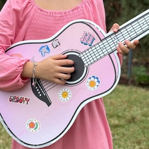 May include: A pink cardboard guitar with various colorful stickers, including a flower with a smiley face, a flower with a blue center, and the text "Good vibes only".