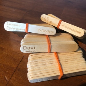May include: A stack of wooden popsicle sticks with personalized messages, including "Luca", "Davi 1 aninho", "Catarina Zimmermann", and "05/01/2010". The sticks are tied together with a rubber band and have the text "@lucachocolaterie" on one of the sticks.