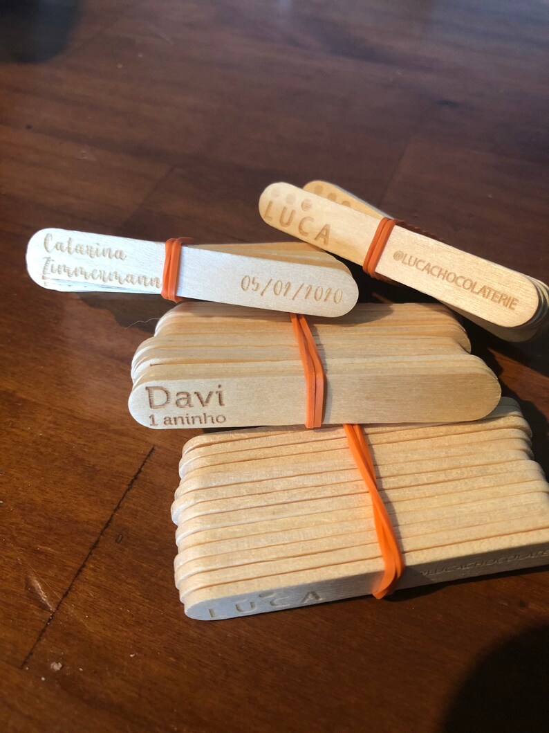 May include: A stack of wooden popsicle sticks with personalized messages, including "Luca", "Davi 1 aninho", "Catarina Zimmermann", and "05/01/2010". The sticks are tied together with a rubber band and have the text "@lucachocolaterie" on one of the sticks.