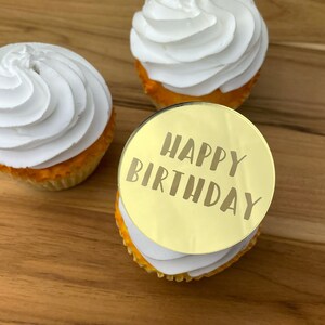 Może przedstawiać: Trzy babeczki z białym kremem. Jedna babeczka ma okrągły złoty topper z napisem "Happy Birthday".
