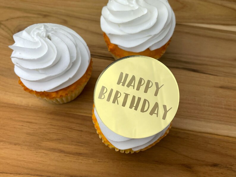 Może przedstawiać: Trzy babeczki z białym kremem. Jedna babeczka ma okrągły złoty topper z napisem "Happy Birthday".