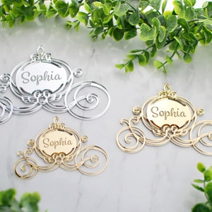 Op de afbeelding: Drie Assepoester-koetsvormige acrylversieringen met de naam "Sophia" gegraveerd op elk. De versieringen zijn in zilver en goud.
