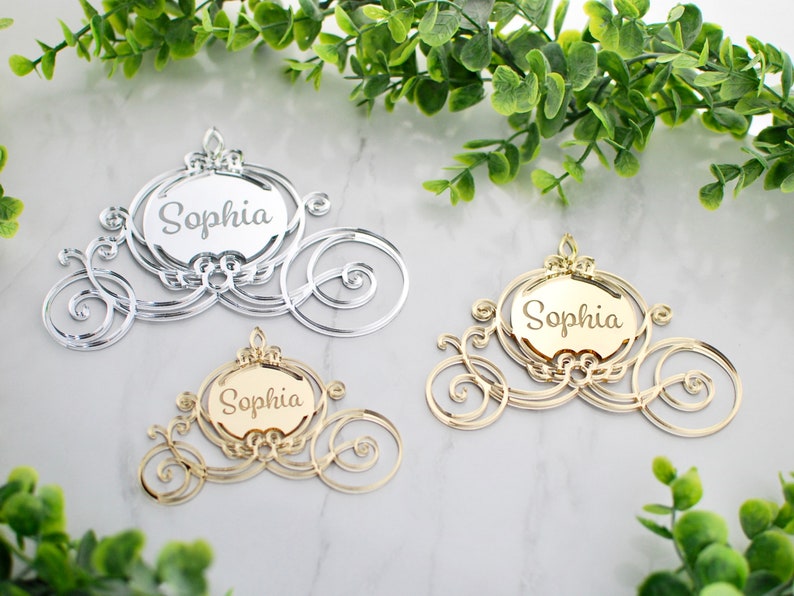 Op de afbeelding: Drie Assepoester-koetsvormige acrylversieringen met de naam "Sophia" gegraveerd op elk. De versieringen zijn in zilver en goud.