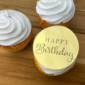 Może przedstawiać: Złoty topper do babeczek z napisem "Happy Birthday" w ozdobnym, pochylonym piśmie. Topper znajduje się na białej babeczce z ż&oacute;łtą polewą.