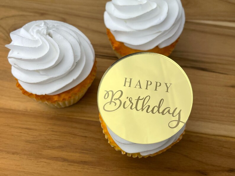 Może przedstawiać: Złoty topper do babeczek z napisem "Happy Birthday" w ozdobnym, pochylonym piśmie. Topper znajduje się na białej babeczce z ż&oacute;łtą polewą.