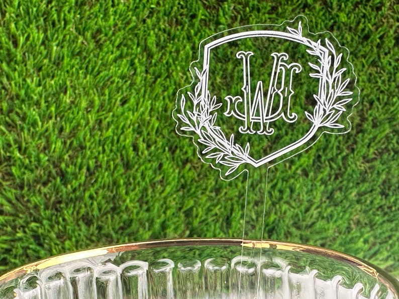 Puede incluir: Un adorno para pastel de acr&iacute;lico transparente con un monograma blanco "WF" dentro de una forma de escudo rodeado de ramas con hojas.