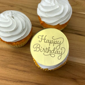 Może przedstawiać: Trzy babeczki z białym kremem. Jedna babeczka ma okrągły złoty topper z napisem "Happy Birthday" w ozdobnym piśmie.