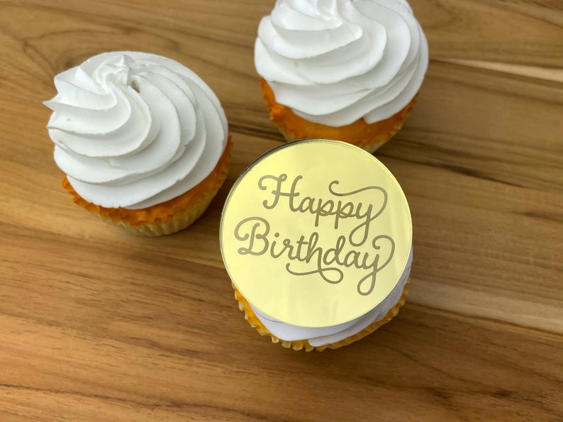 Może przedstawiać: Trzy babeczki z białym kremem. Jedna babeczka ma okrągły złoty topper z napisem "Happy Birthday" w ozdobnym piśmie.