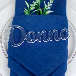 May include: A clear acrylic place card with the name "Donna" written in a cursive font. The place card is sitting on a blue napkin.