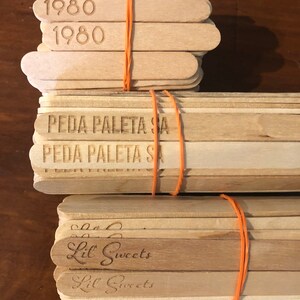 May include: A stack of wooden popsicle sticks with the text "1980", "PEDA PALETA SA", and "Lil Sweets" printed on them. The sticks are tied together with a rubber band.