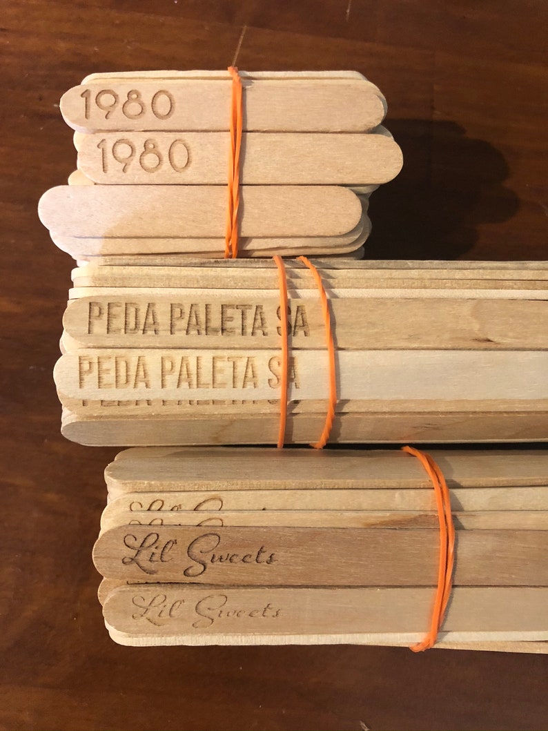 May include: A stack of wooden popsicle sticks with the text "1980", "PEDA PALETA SA", and "Lil Sweets" printed on them. The sticks are tied together with a rubber band.