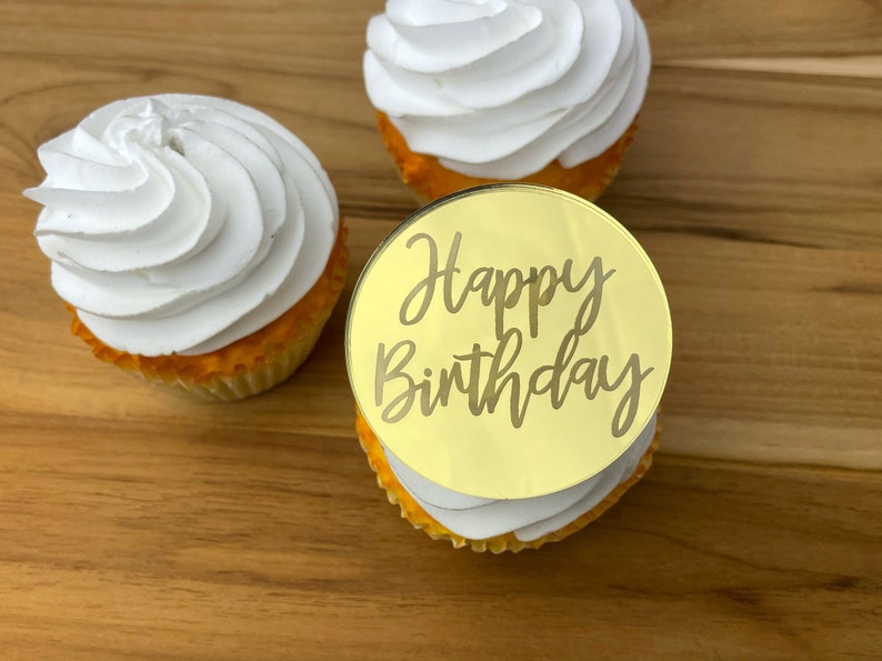 Może przedstawiać: Złoty topper do babeczek z napisem "Happy Birthday" na białej babeczce z białym lukrem.