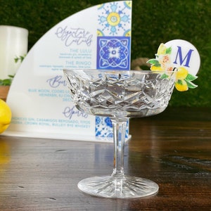 May include: A clear glass coupe champagne glass with a decorative lemon and floral design drink marker with the letter "M" on a wooden table. The glass is empty. The drink marker is on the rim of the glass.