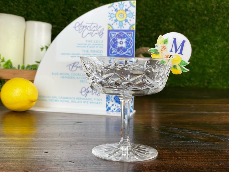May include: A clear glass coupe champagne glass with a decorative lemon and floral design drink marker with the letter "M" on a wooden table. The glass is empty. The drink marker is on the rim of the glass.