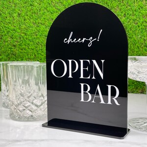 May include: Black acrylic Open Bar sign with a rounded top, featuring the word "cheers!" in script and "OPEN BAR" in bold, white capital letters. The sign is displayed on a white surface, with clear glassware in the background. Perfect for a wedding or party.