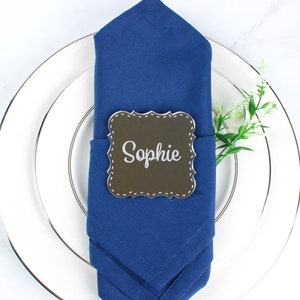 May include: A white dinner plate with a blue napkin folded into a triangular shape. The napkin has a brown place card with the name "Sophie" written in white. The plate is set with a silver fork and knife.
