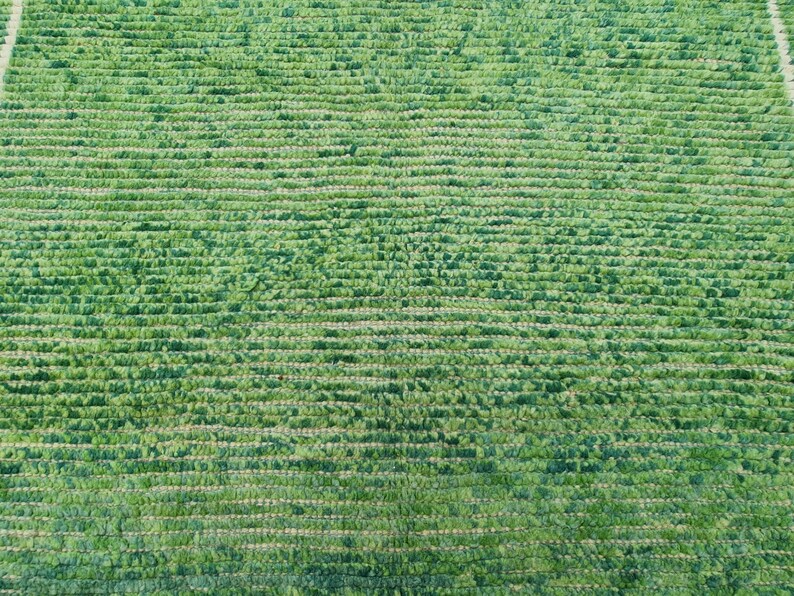 Puede incluir: Primer plano de una alfombra verde con un patr&oacute;n horizontal texturizado. La alfombra tiene un pelo denso y rizado, creando una apariencia suave y afelpada. El color es un verde vibrante y natural, lo que sugiere un producto hecho a mano o de fibra natural.