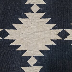 Puede incluir: Primer plano de una alfombra tejida con un patr&oacute;n geom&eacute;trico. La alfombra presenta un dise&ntilde;o repetitivo de diamantes y tri&aacute;ngulos en una combinaci&oacute;n de colores contrastantes azul marino y blanco roto. La textura de la alfombra es visible.