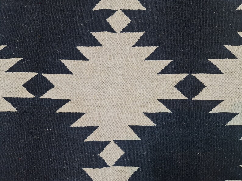 Puede incluir: Primer plano de una alfombra tejida con un patr&oacute;n geom&eacute;trico. La alfombra presenta un dise&ntilde;o repetitivo de diamantes y tri&aacute;ngulos en una combinaci&oacute;n de colores contrastantes azul marino y blanco roto. La textura de la alfombra es visible.