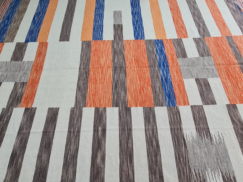 May include: Geometric patterned fabric featuring vertical stripes in shades of orange, blue, brown, and gray against a white background. The design has a modern, abstract aesthetic, suitable for home decor or textile projects.