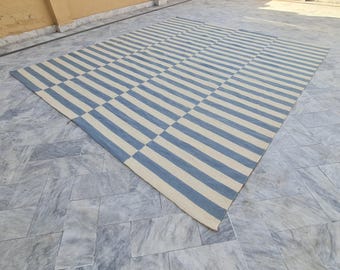 May include: A large, rectangular rug with a geometric pattern of alternating blue and off-white stripes. The rug is made of a woven material and is laid out on a tiled surface. The rug's design creates a visually appealing, modern aesthetic.