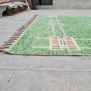 Puede incluir: Una alfombra rectangular verde con un dise&ntilde;o geom&eacute;trico. La alfombra presenta un fondo verde claro con un patr&oacute;n de l&iacute;neas blancas y formas rectangulares en tonos naranja, azul y crema. Tiene una franja en un lado.