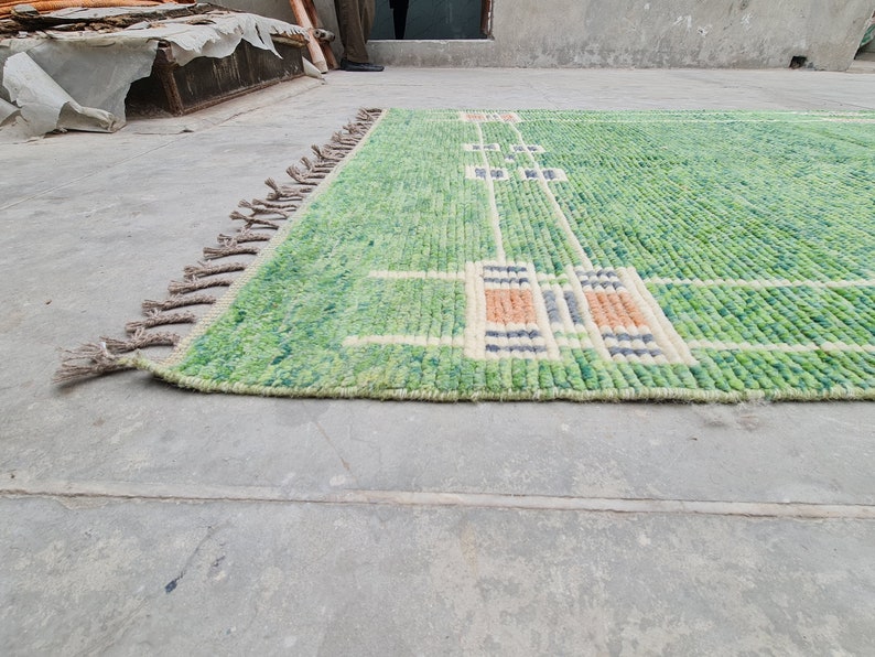 Puede incluir: Una alfombra rectangular verde con un dise&ntilde;o geom&eacute;trico. La alfombra presenta un fondo verde claro con un patr&oacute;n de l&iacute;neas blancas y formas rectangulares en tonos naranja, azul y crema. Tiene una franja en un lado.