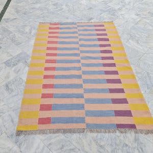 May include: Rectangular area rug with a geometric pattern. The rug features horizontal stripes in shades of yellow, pink, blue, red, and purple. The rug has a fringed edge. The rug is designed for indoor use.