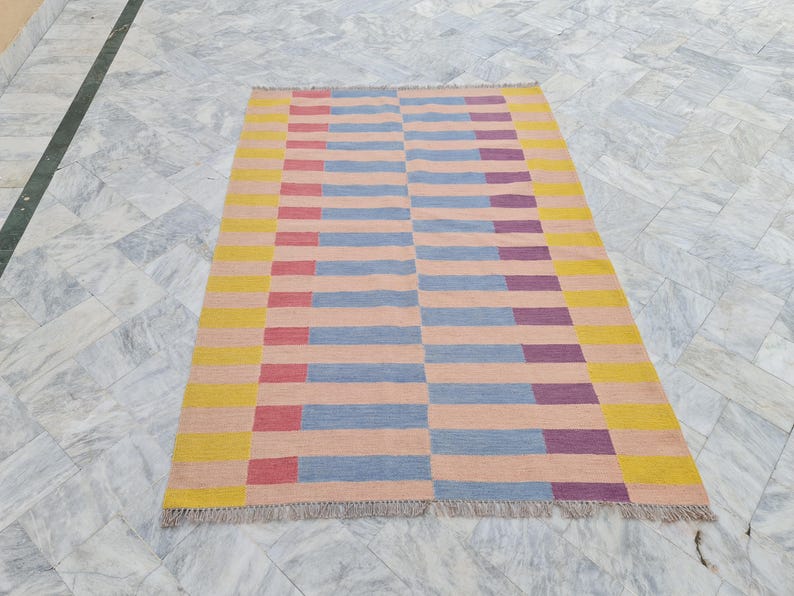 May include: Rectangular area rug with a geometric pattern. The rug features horizontal stripes in shades of yellow, pink, blue, red, and purple. The rug has a fringed edge. The rug is designed for indoor use.